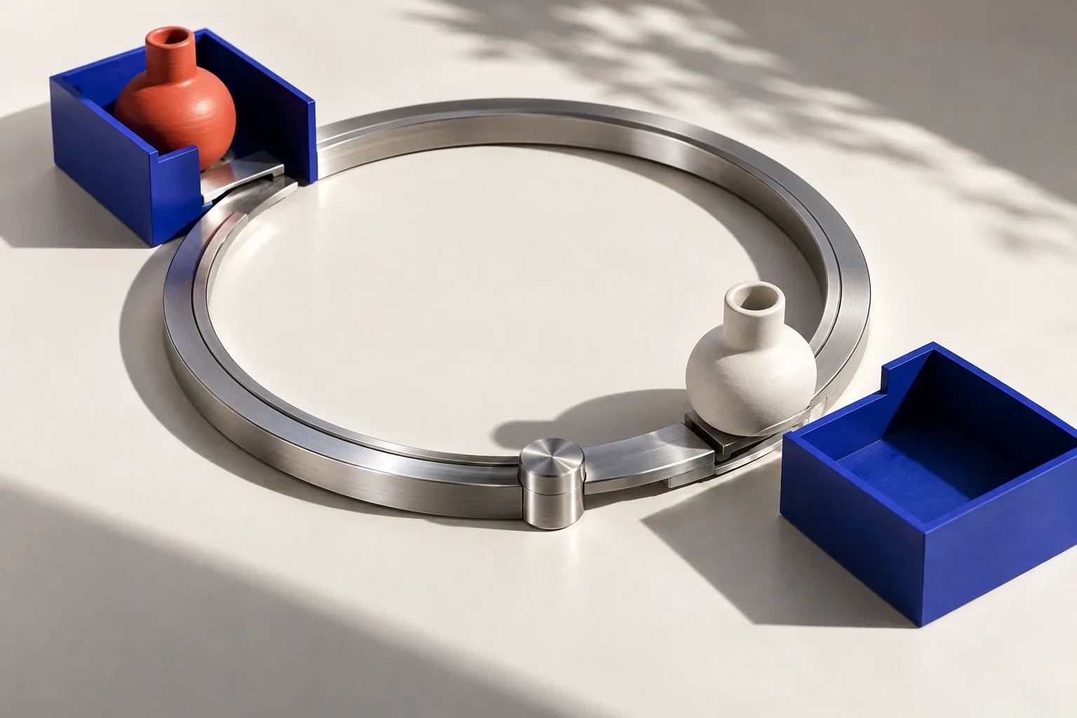 A circular aluminium return track exchanges two ceramic objects between cobalt trays.
